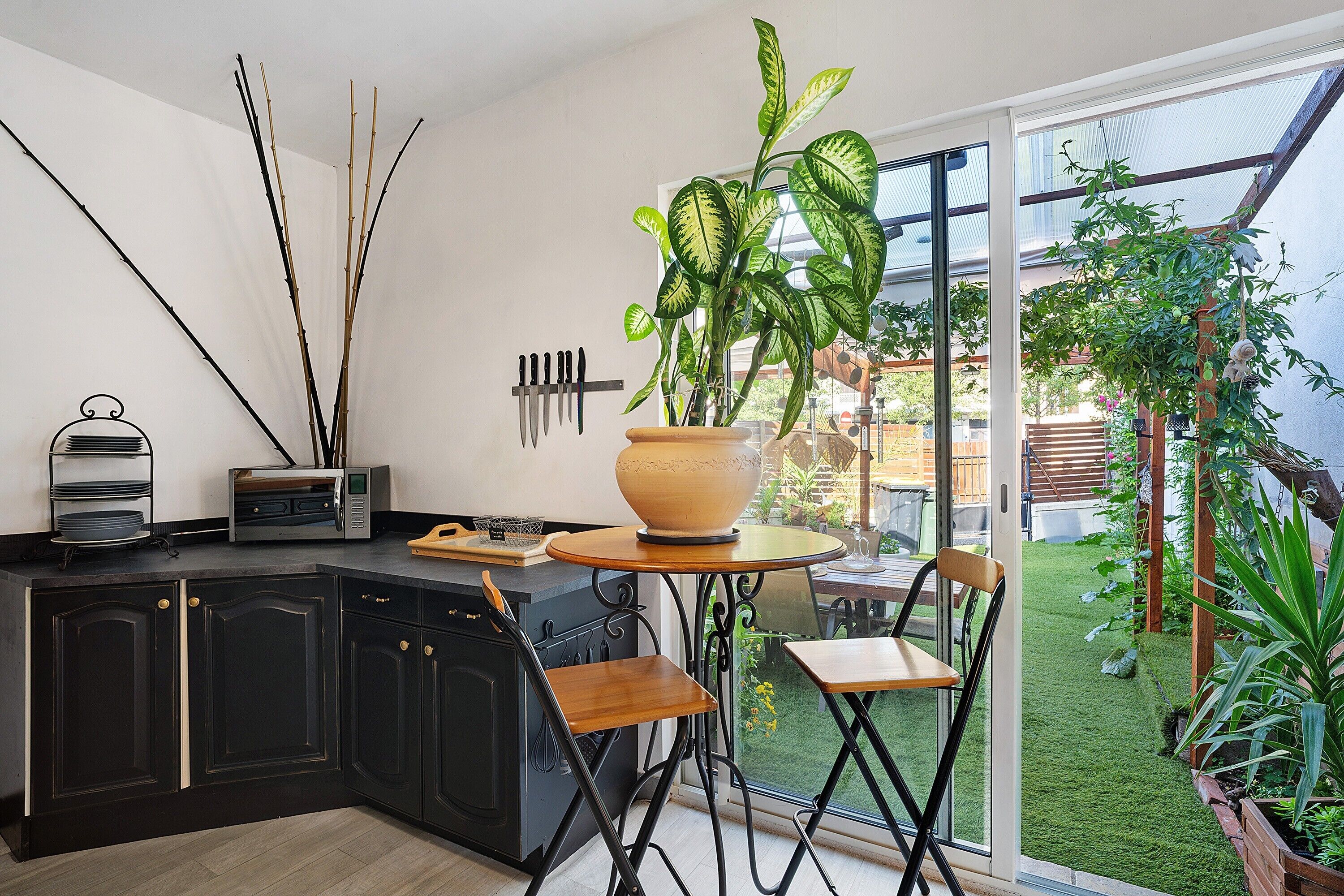 A kitchen with dark cabinetry, a dining area with a table and chairs, a large potted plant, and a view of a garden through a glass door.
