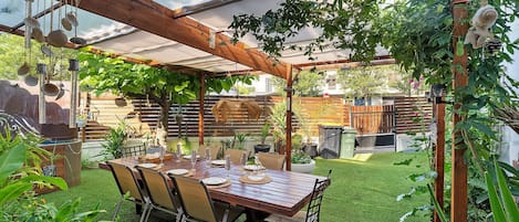 A covered patio with a wooden table set for dining, surrounded by chairs and potted plants.