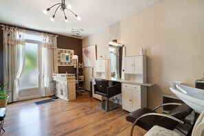 Een kapsalon met een houten vloer, een zwarte kappersstoel, een wit bureau en een grote spiegel.