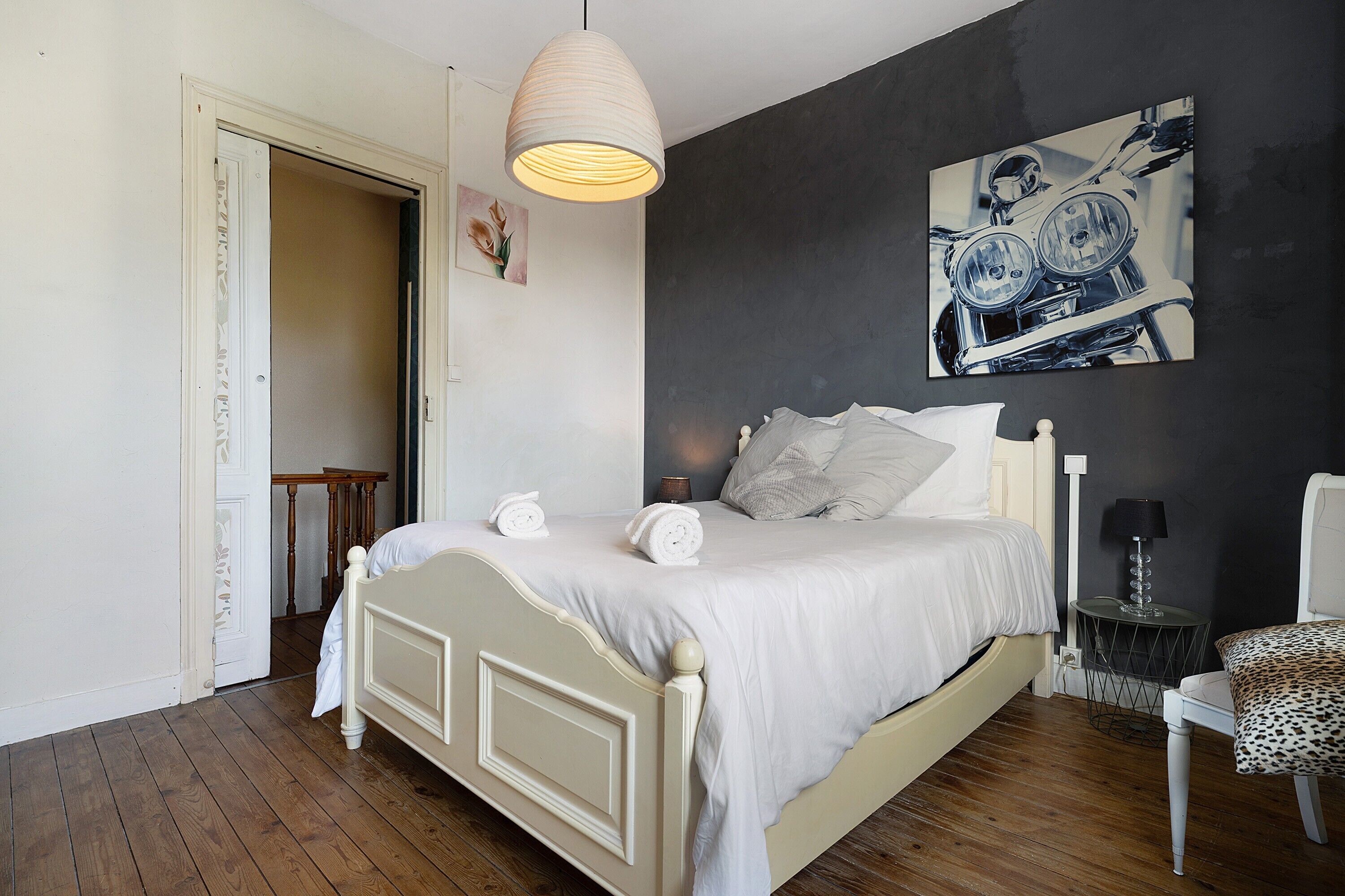 A bedroom with a bed, a hanging light fixture, a wall-mounted painting, and a chair.