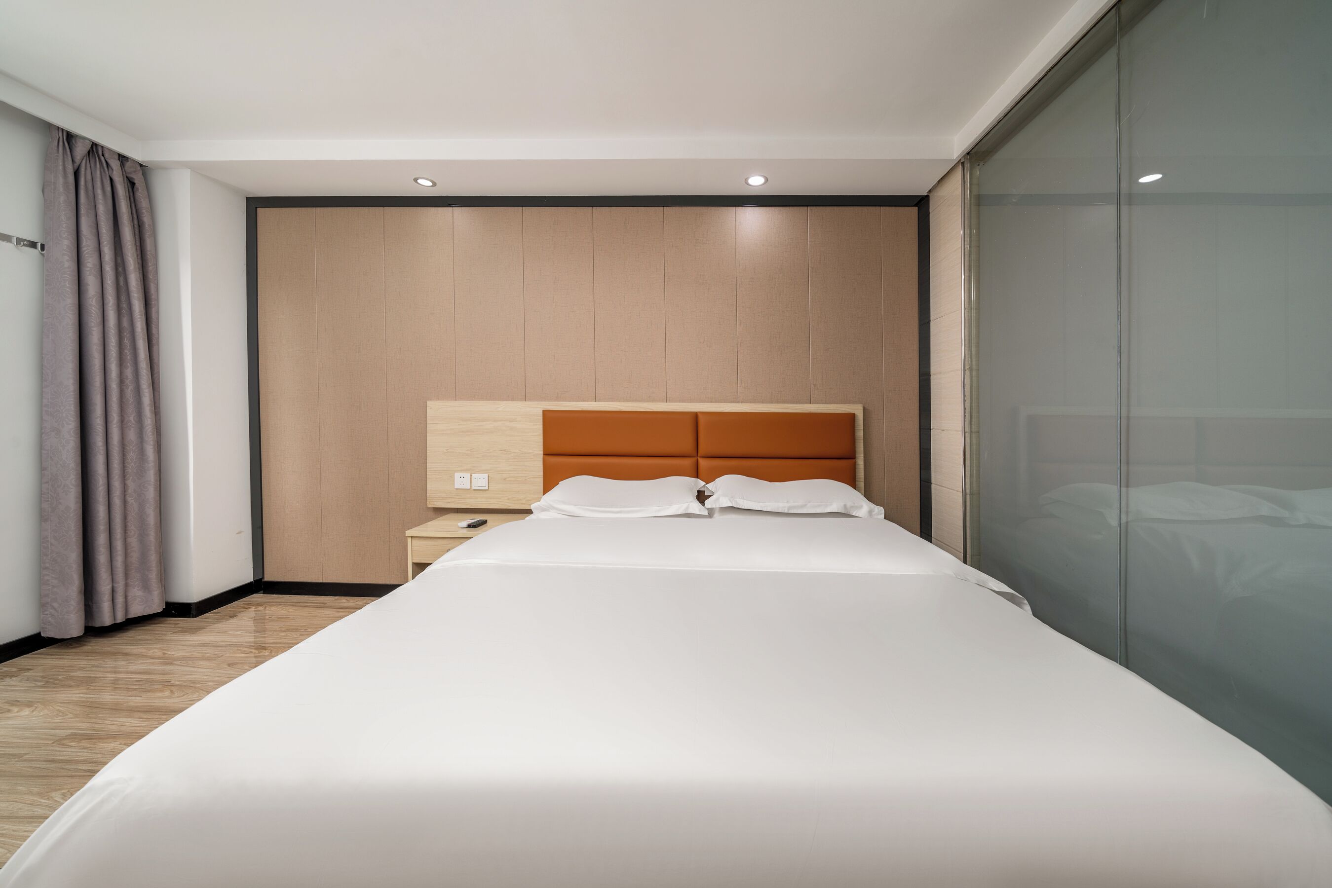 A modern hotel room with a large bed, wooden headboard, and a glass partition wall.