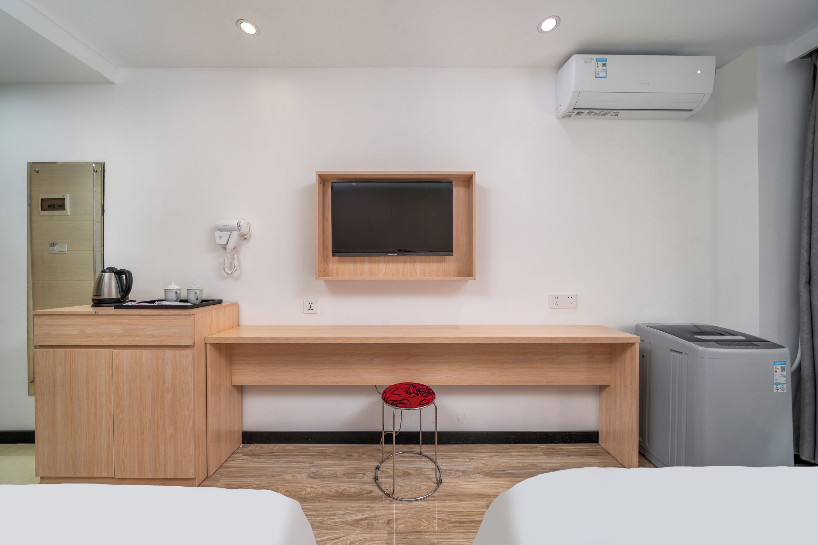 A hotel room with a wooden desk, a TV mounted on the wall, a small stool, a trash bin, and a coffee maker.