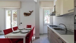 A modern kitchen with white cabinets, a stainless steel sink, and a dining area with red chairs and a white table. - Hostdomus - Turtle House (Borgio)