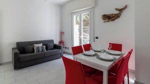 A dining area with a white table and red chairs, a gray sofa, and a ceiling fan. - Hostdomus - Turtle House (Borgio)