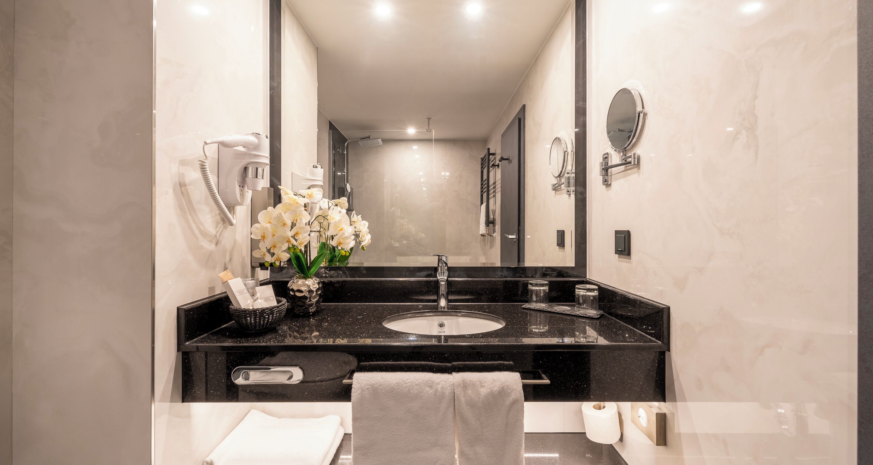 A modern bathroom with a black marble sink, a mirror, a towel rack, and a vase of white flowers.