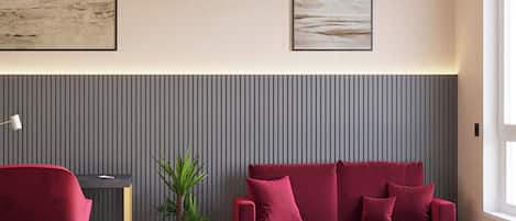 Een moderne kamer met een rode bank, een klein rond tafeltje, een rode bureaustoel, een potplant en twee kunstwerken in lijsten aan de muur.