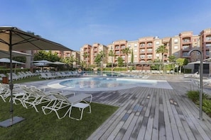 A pool area with lounge chairs, a wooden deck, and a row of apartment buildings in the background. - Casa Arte-Luxury Apartments Islantilla-RuitenAppartementen (Lepe)