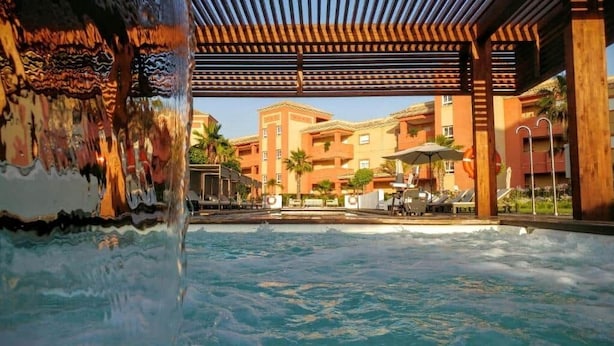 A pool area with a wooden pergola, a water feature, and a view of multi-story buildings with balconies.