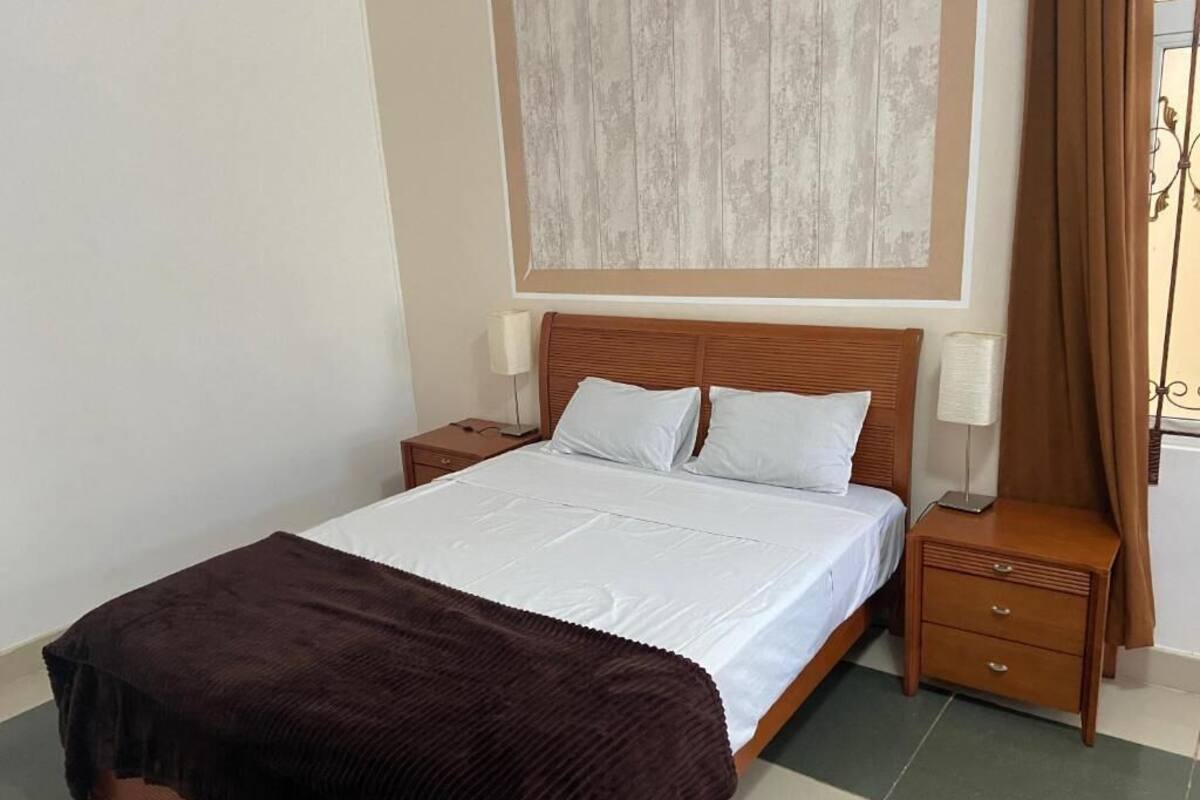 A neatly made bed with a brown blanket, a wooden headboard, a bedside table with a lamp, and a wall-mounted artwork.