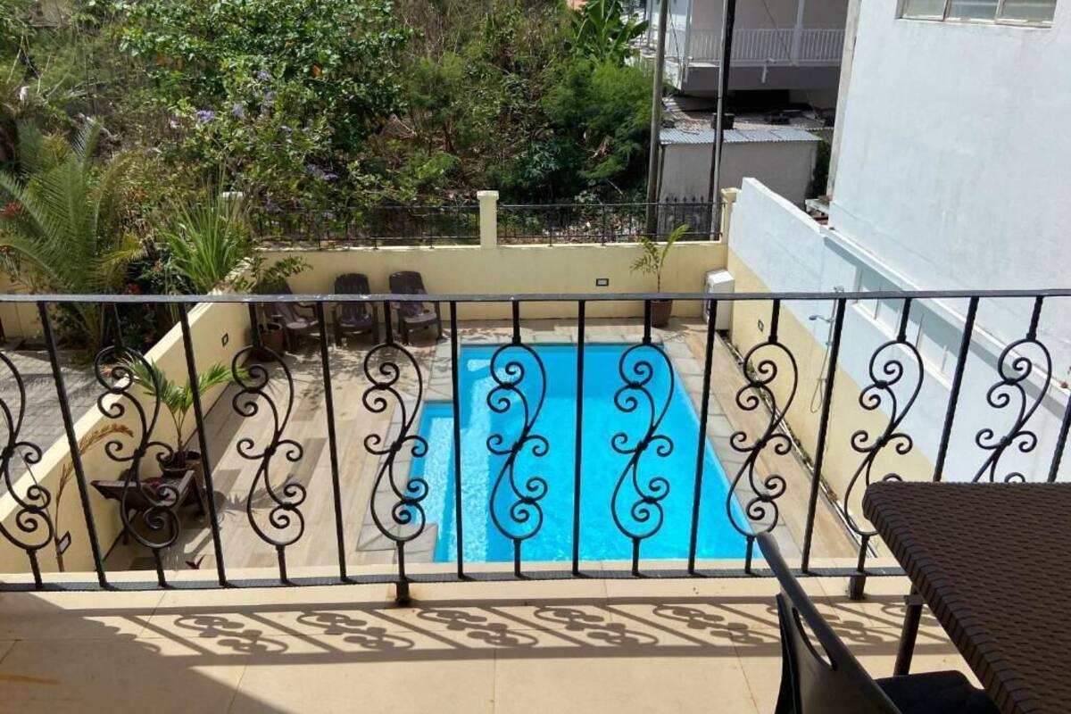 A balcony with a pool, surrounded by a metal railing, and overlooking a residential area with trees and buildings.
