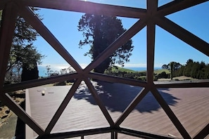 Une vue à travers une structure géométrique dotée d’une terrasse en bois, donnant sur un paysage composé d’arbres et de collines.