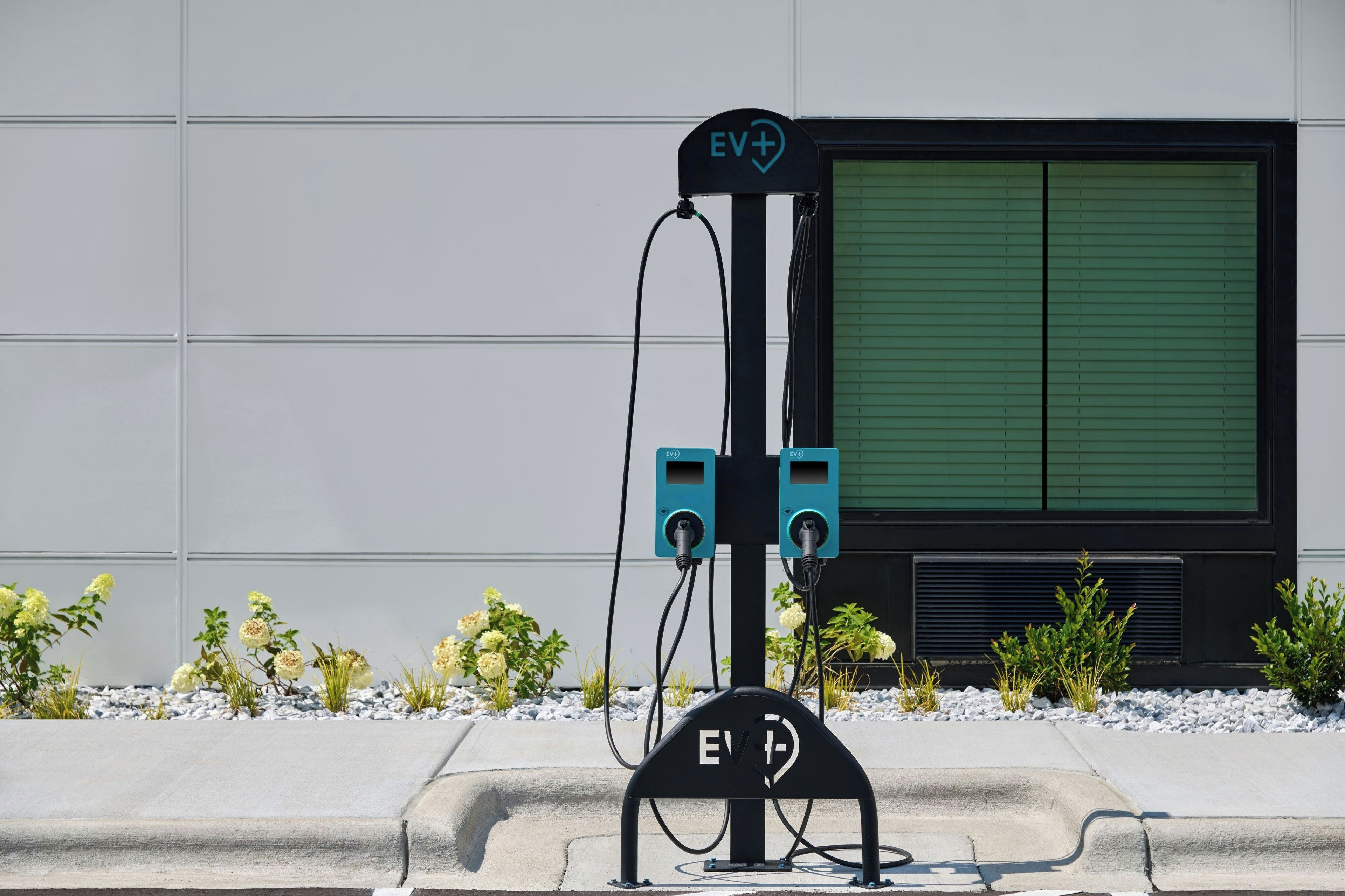 An EV charging station with a sign, a green shuttered window, and a concrete curb with plants.