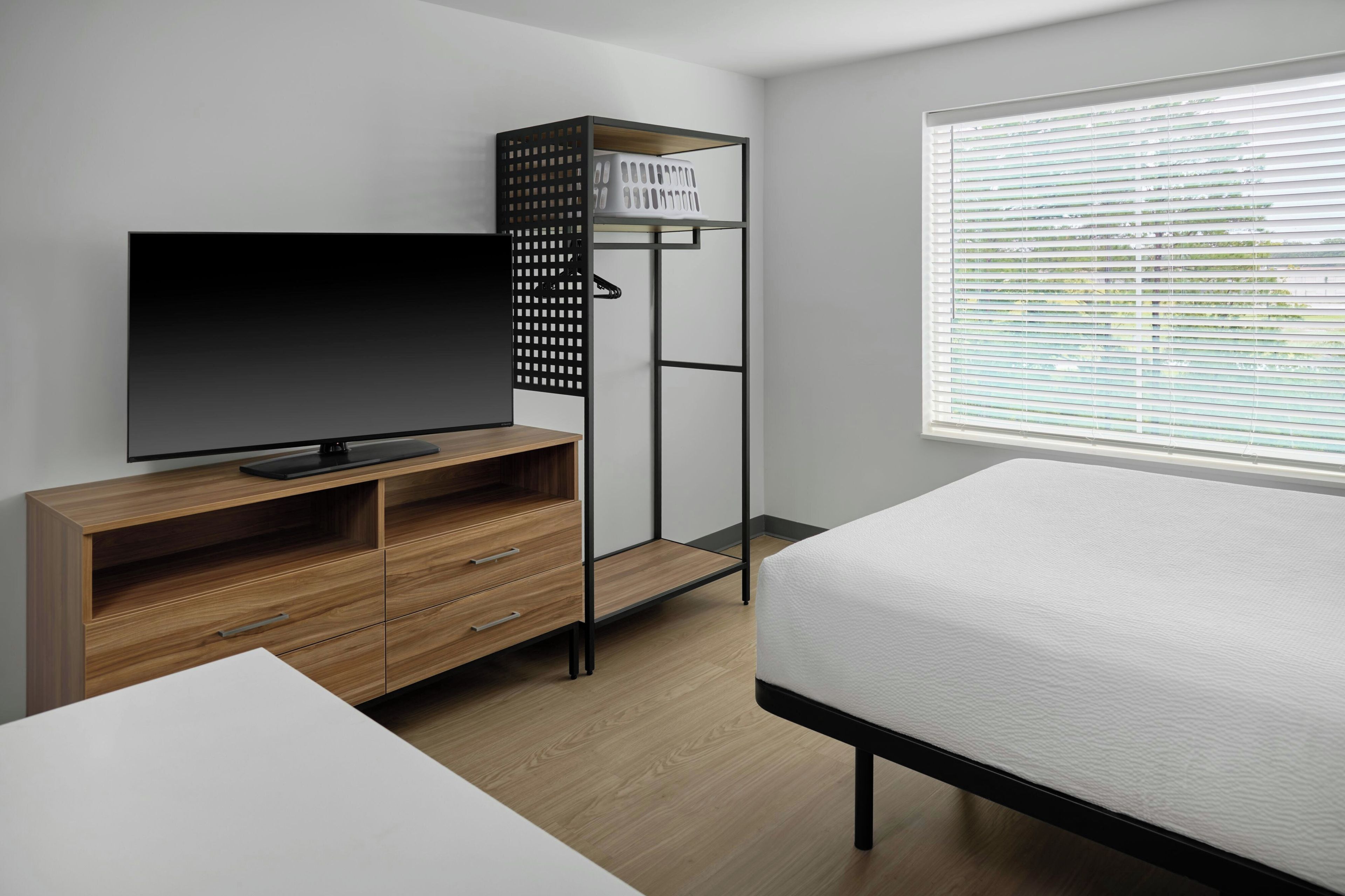 A modern bedroom with a wooden TV stand, a bed, a built-in wardrobe, and a window with blinds.