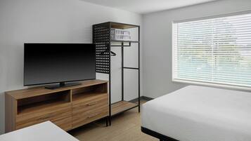 A modern bedroom with a wooden TV stand, a bed, a built-in wardrobe, and a window with blinds.
