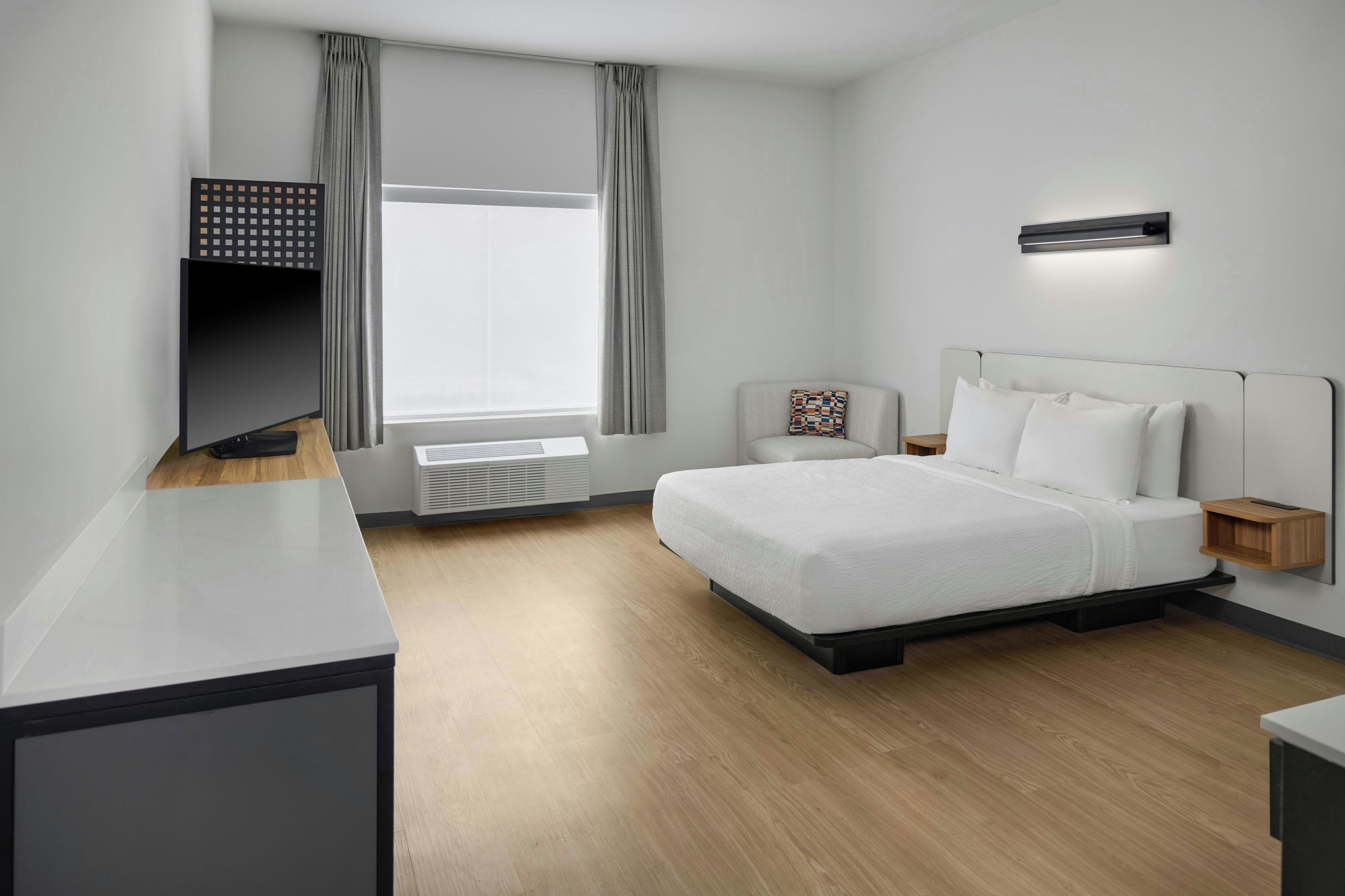 A modern hotel room with a bed, a desk, a chair, a wall-mounted air conditioner, and a window with curtains.