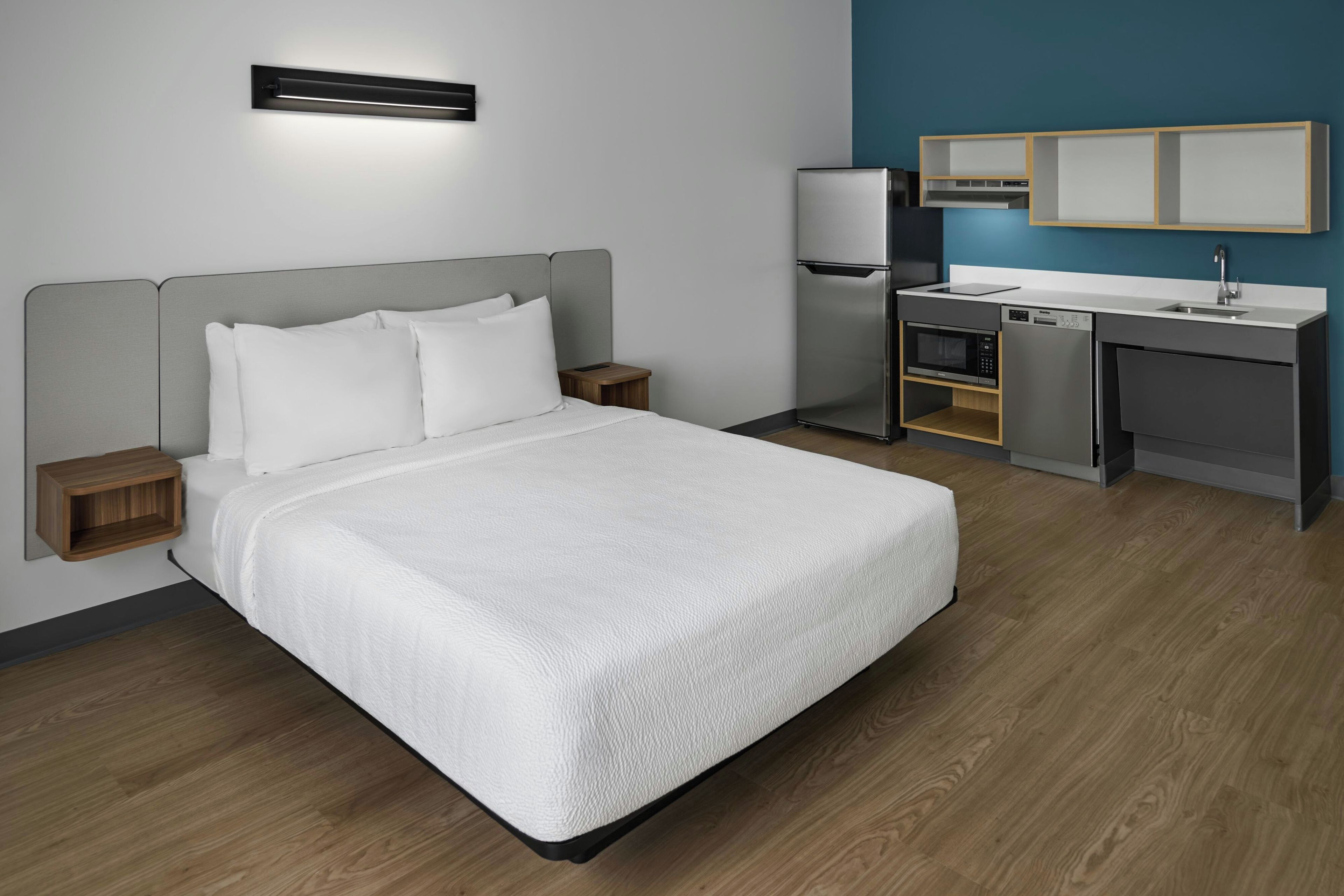 A hotel room with a bed, bedside tables, a desk with a microwave, a refrigerator, and a built-in oven.
