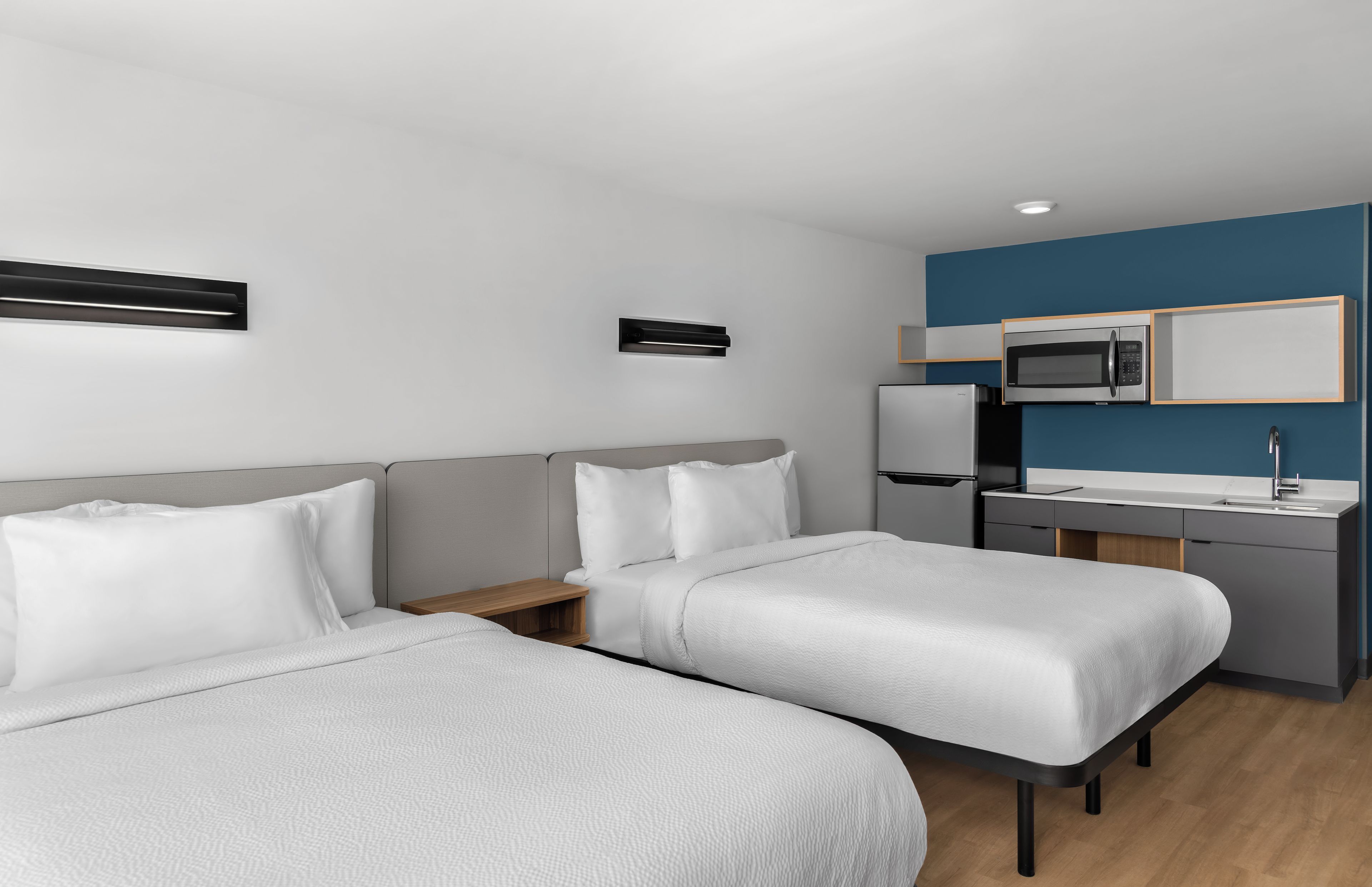 Quarto de hotel moderno com duas camas de solteiro, uma mesa com micro-ondas e uma pia pequena.