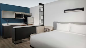A modern hotel room with a bed, a desk with a microwave, a small kitchenette, and a wall-mounted light fixture. - StudioRes by Marriott Wilmington (Wilmington)