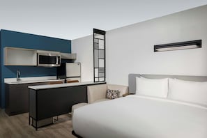 A modern hotel room with a bed, a desk with a microwave, a small kitchenette, and a wall-mounted light fixture. - StudioRes by Marriott Wilmington (Wilmington)