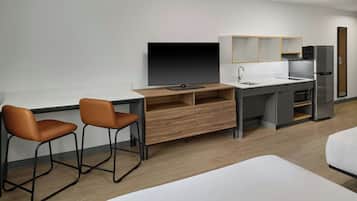 A modern hotel room with a wooden desk, two chairs, a flat-screen TV, a mini-fridge, a sink, and a bed.