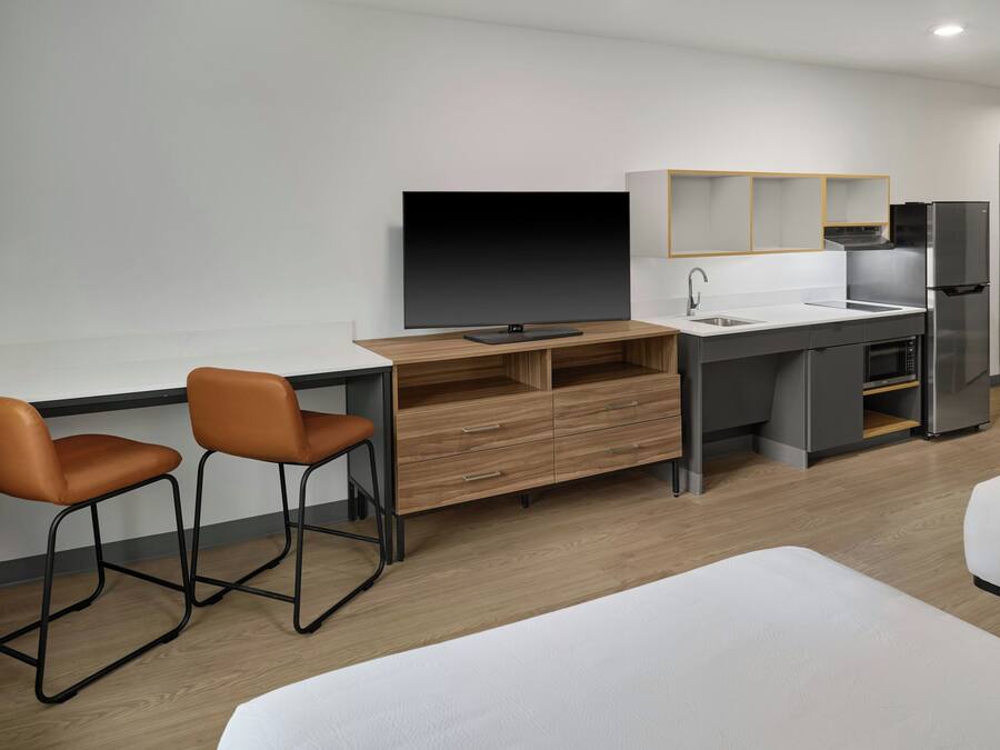 Una habitación de hotel moderna con un escritorio de madera, dos sillas, un televisor de pantalla plana, un mini refrigerador, un lavamanos y una cama.