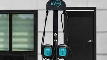 An EV+ charging station with two charging points, mounted on a pole beside a building with a window and a door.