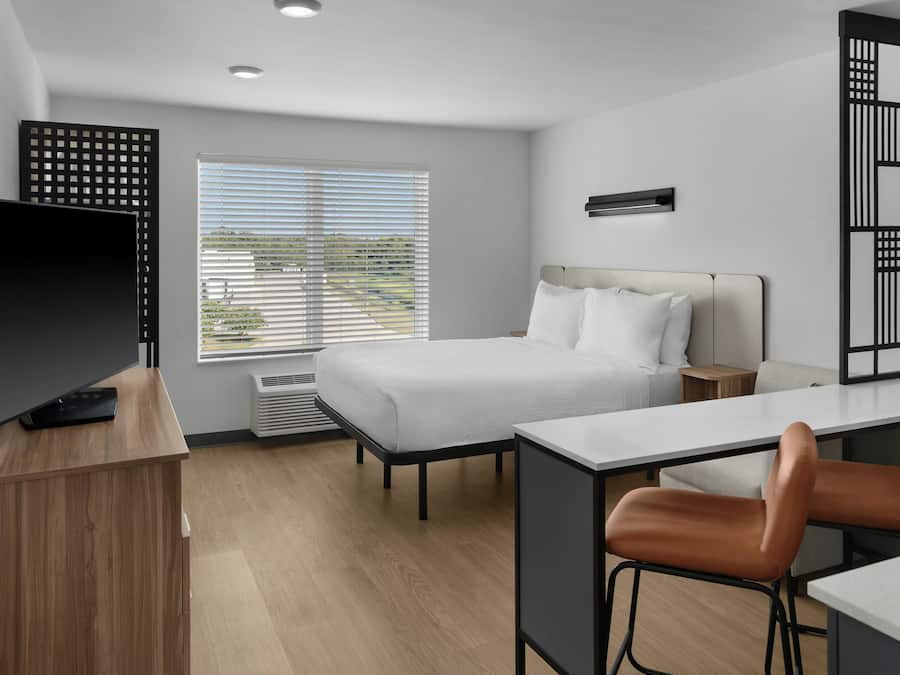 Habitación de hotel moderna con cama, escritorio, silla, televisor y ventana con persianas.