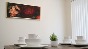 Uma sala de jantar com uma mesa posta para quatro pessoas, uma pequena planta em vaso e um quadro com flores na parede.