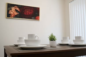 Un comedor con una mesa dispuesta para cuatro, una pequeña planta en maceta y un cuadro de flores en la pared.