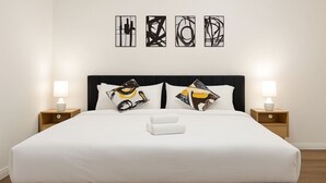 Cama bem arrumada com lençóis brancos, dois travesseiros com desenhos abstratos e uma toalha branca. Duas luminárias de cabeceira estão sobre mesas de cabeceira de madeira.