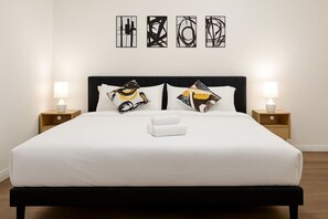 Una cama bien hecha con sábanas blancas, dos almohadas con diseños abstractos y una toalla blanca. Hay dos lámparas de noche sobre mesitas de madera.