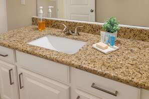 Un baño con encimera de granito, un lavabo blanco, un espejo, una planta en maceta y artículos de aseo.