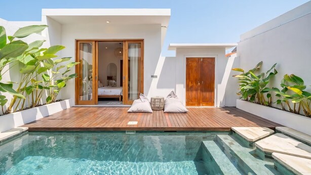 A modern poolside area with a wooden deck, a glass-enclosed bedroom, and a white exterior wall. - Sunset Heaven C6 Villa (Denpasar)