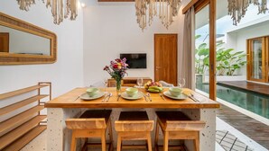 A dining area with a wooden table, chairs, a vase of flowers, and a television on the wall. - Sunset Heaven C6 Villa (Denpasar)