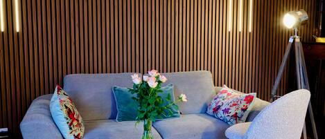 A modern living room with a sofa, a chair, a glass coffee table, a vase with flowers, and a lamp.