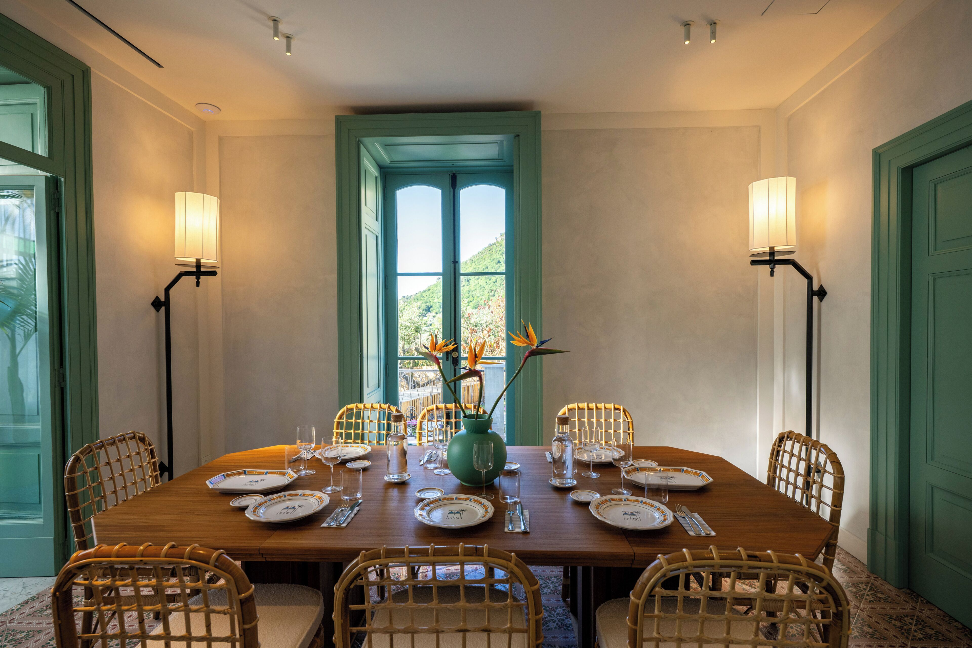 A dining room with a wooden table set for four, surrounded by wicker chairs, with a vase of flowers on the table.