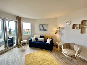 Ein modernes Wohnzimmer mit einem blauen Sofa, einem beigen Sessel, einem gelben Teppich und einem großen Fenster mit Blick.