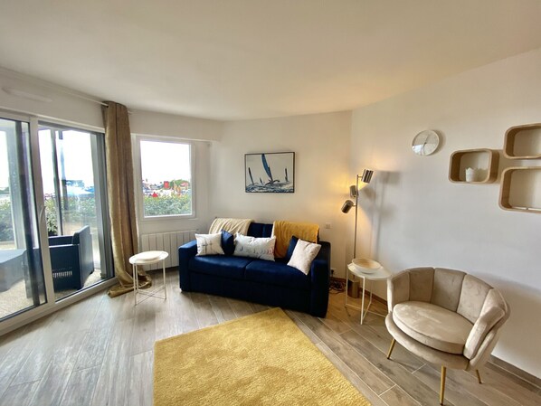 Een moderne woonkamer met een blauwe bank, een beige fauteuil, een geel vloerkleed en een groot raam met uitzicht.