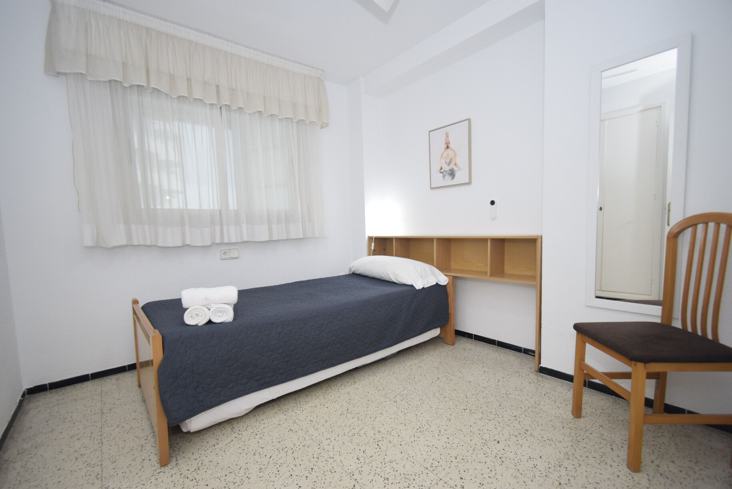 A single bed with a headboard, a chair, a window with sheer curtains, and a framed picture on the wall.