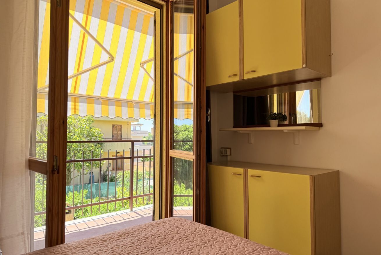 Une pièce avec un lit, des armoires jaunes, une petite étagère et une vue sur un balcon doté d’un auvent à rayures.