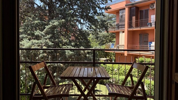 Un balcon avec une table en bois et deux chaises, donnant sur un immeuble doté de balcons et sur un jardin.