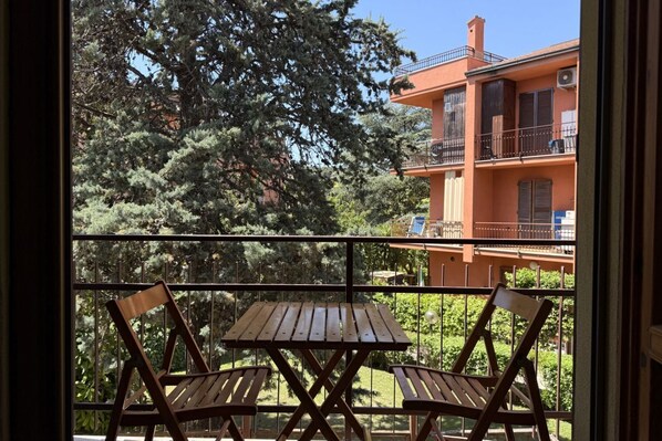 Ein Balkon mit Holztisch und zwei Stühlen, mit Blick auf ein Gebäude mit Balkonen und einem Garten.