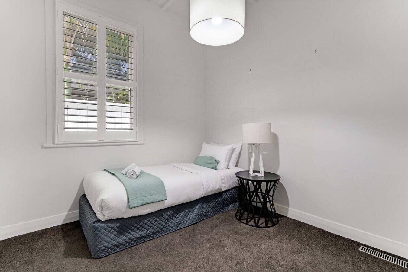 A bedroom with a bed, a bedside table with a lamp, a window with blinds, and a carpeted floor.
