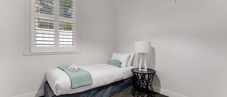 A bedroom with a bed, a bedside table with a lamp, a window with blinds, and a carpeted floor.