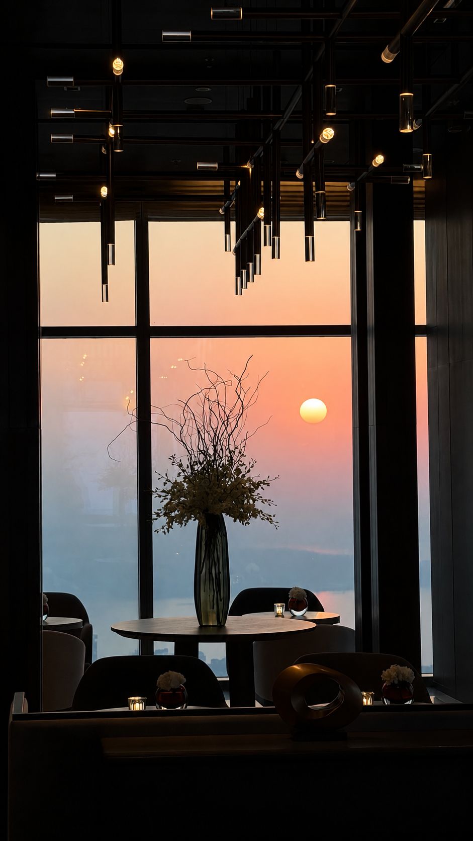 A dining area with a table and chairs, a vase with flowers, and a view of the sunset through large windows.