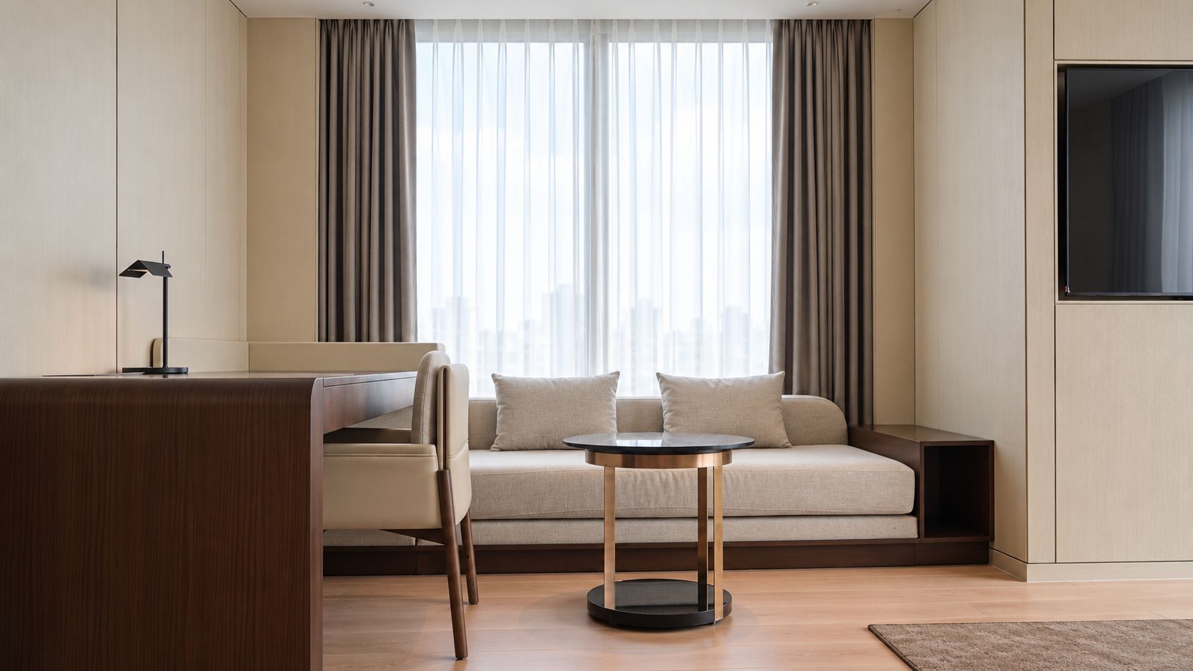 A modern hotel room with a sofa, a small round table, and a desk with a lamp.