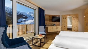 Een moderne hotelkamer met een groot raam, een flatscreen-tv, een houten bureau, een bank en een balkon met uitzicht op besneeuwde bergen.