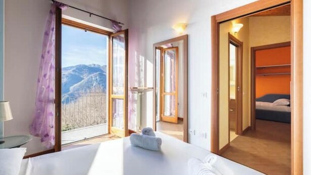 Een hotelkamer met een bed, een raam met uitzicht op bergen, een badkamer met douche, en een deur die naar een andere kamer leidt.