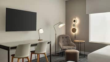 Habitación de hotel con escritorio, dos sillas, un sofá, televisión, lámpara de pie y luz empotrada en la pared.