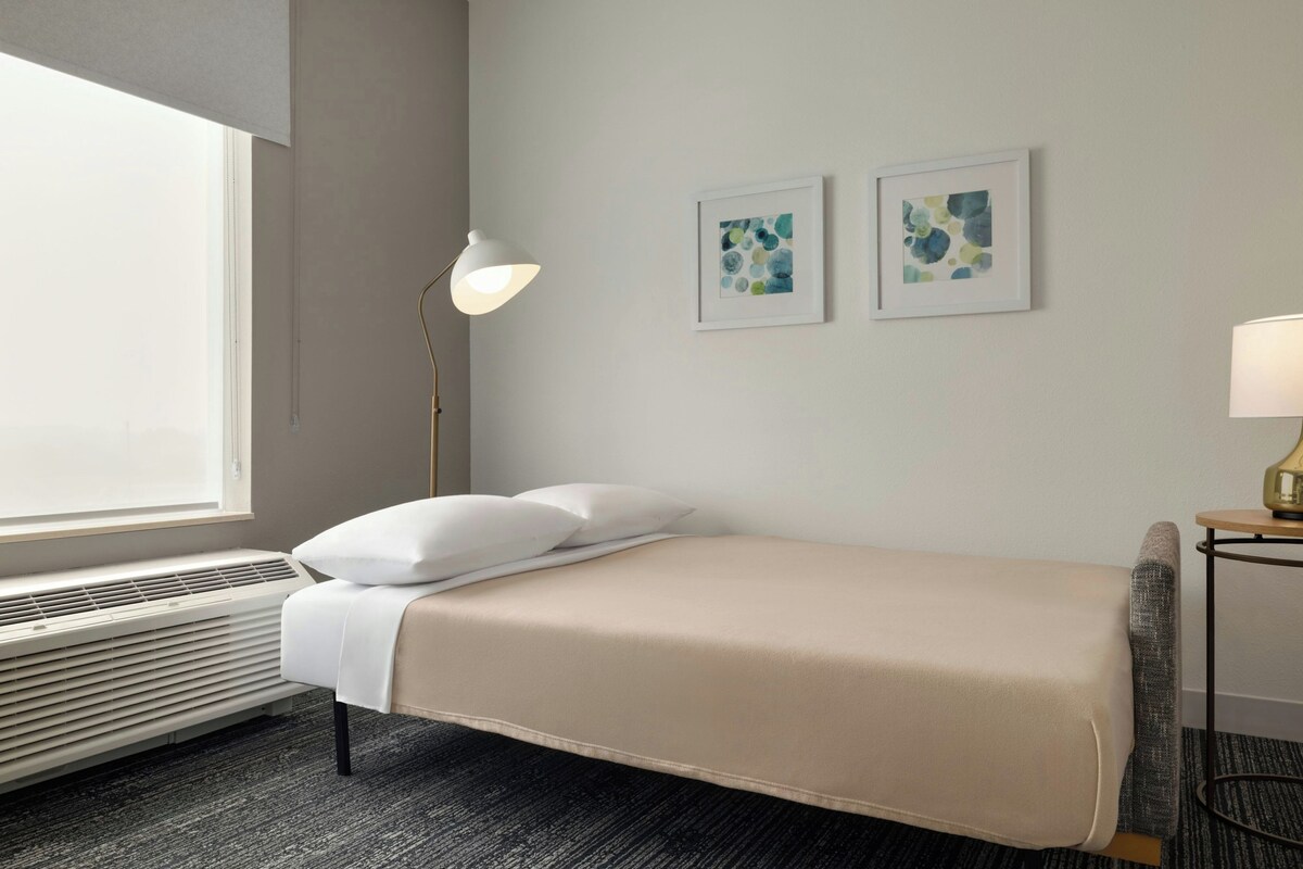 A hotel room with a single bed, a standing lamp, two wall art pieces, and a window with blinds.